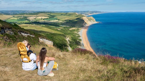 A woman and a man with a baby carrier looking out over the Jurassic Coast from Golden Cap in Dorset, on a summer's day